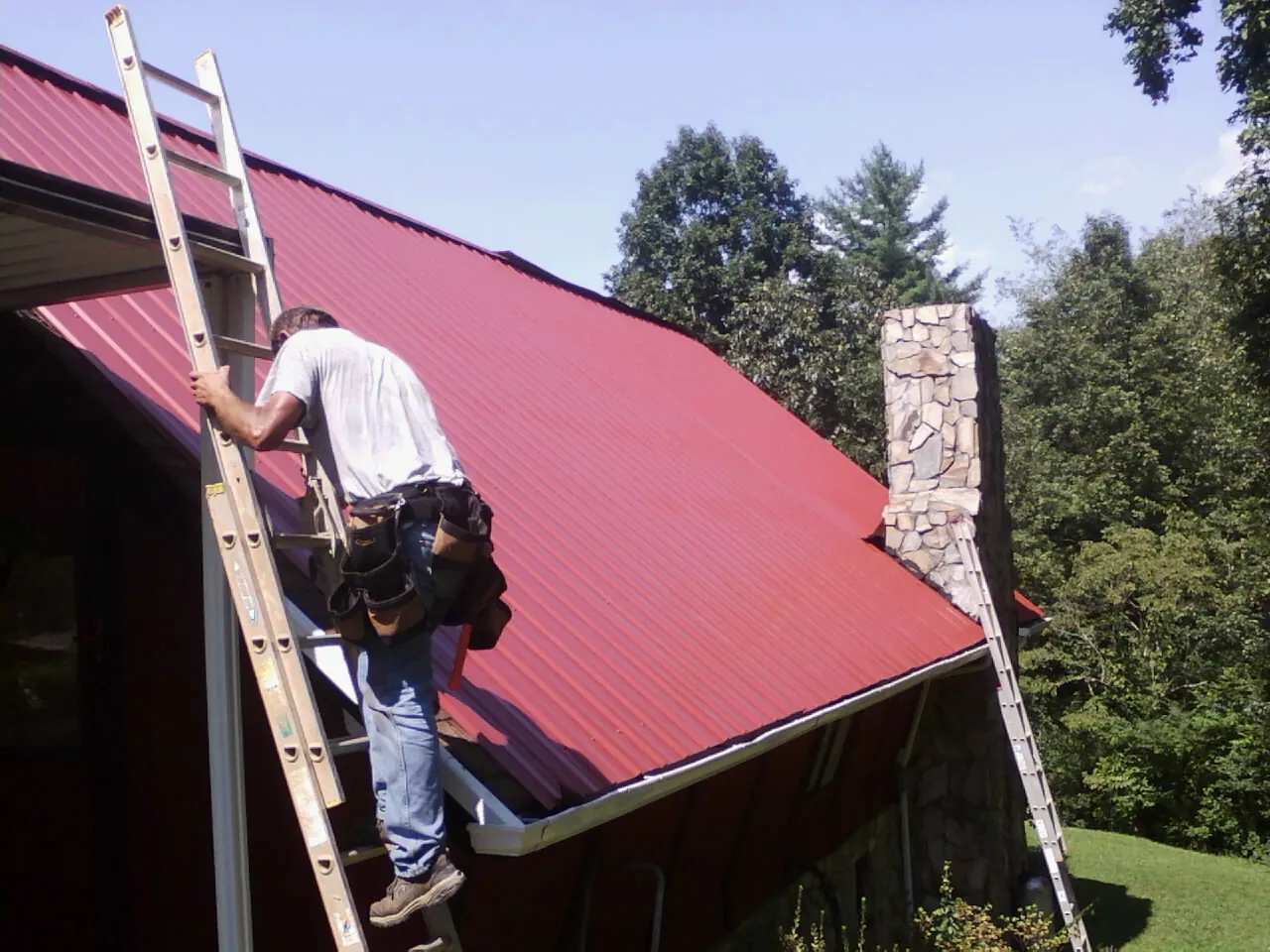 Roofer on a ladder beside a red metal roof and stone chimney — Soffit & Fascia Repair by Roofing Brothers in Coyle